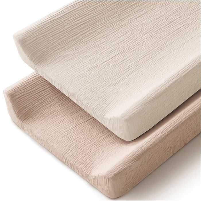 Two changing pad covers stacked at an angle on a white background. The top cover is ivory/cream and the bottom is a warm clay/blush tone. Both feature a crinkled muslin texture with softly rounded corners.