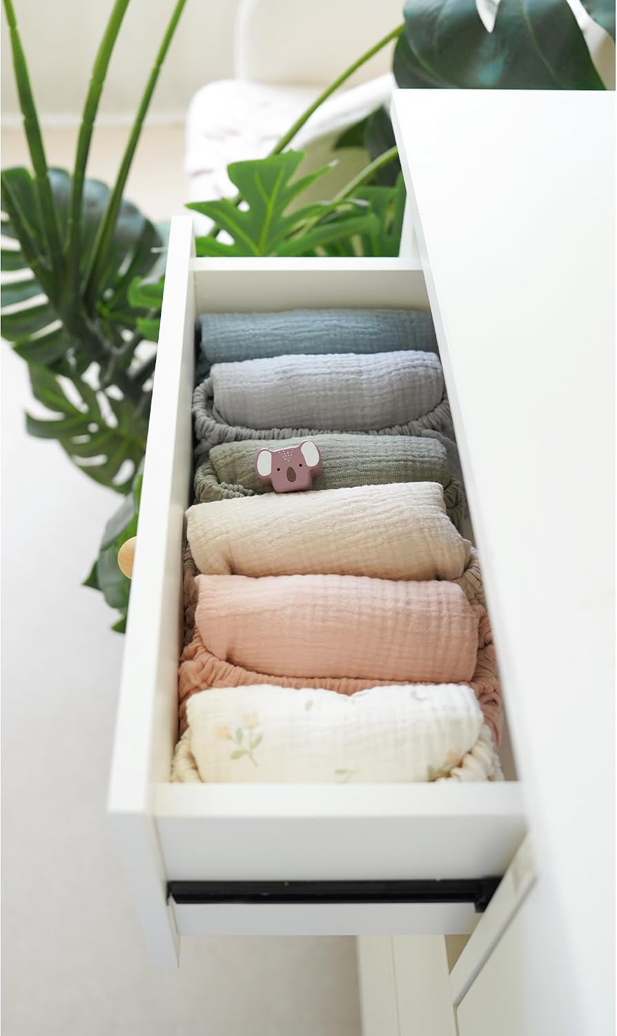 An open white dresser drawer neatly organized with rolled muslin cloths in soft tones: dusty blue, sage green, gray, cream, blush pink, and off-white with a subtle floral print. A small koala figurine is tucked among the rolls.