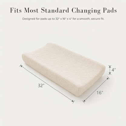 Changing pad cover in cream/off-white textured muslin fabric shown on a contoured pad from a three-quarter angle. Dimension arrows indicate 32" long x 16" wide x 4" thick. Text reads 'Fits Most Standard Changing Pads.'