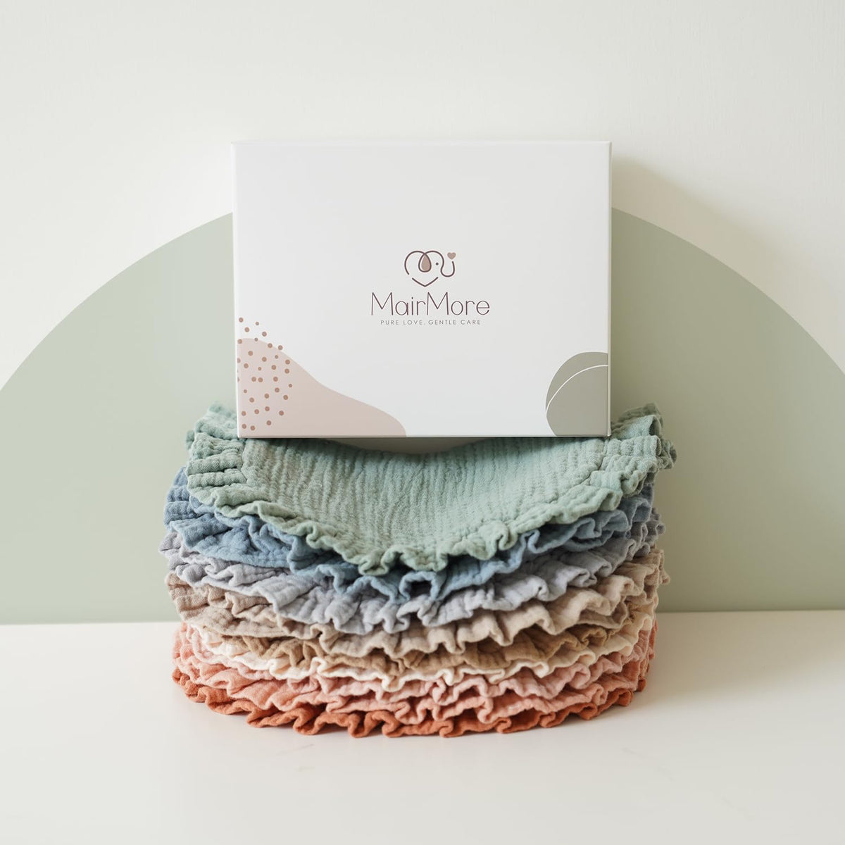 Stack of MairMore muslin bandana bibs in assorted muted tones -- sage green, blue, gray, beige, pink, and terracotta -- with ruffled edges, topped by a white branded gift box reading 'MairMore, Pure Love, Gentle Care'.