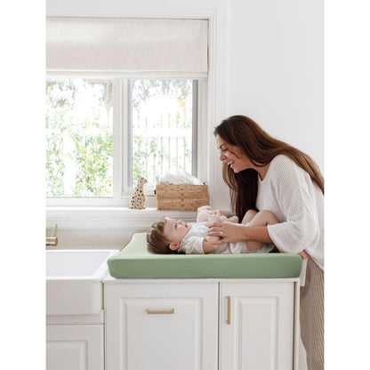 A smiling woman leans over a baby lying on a changing pad covered in a soft sage green textured cover. The pad sits atop white cabinetry near a bright window.