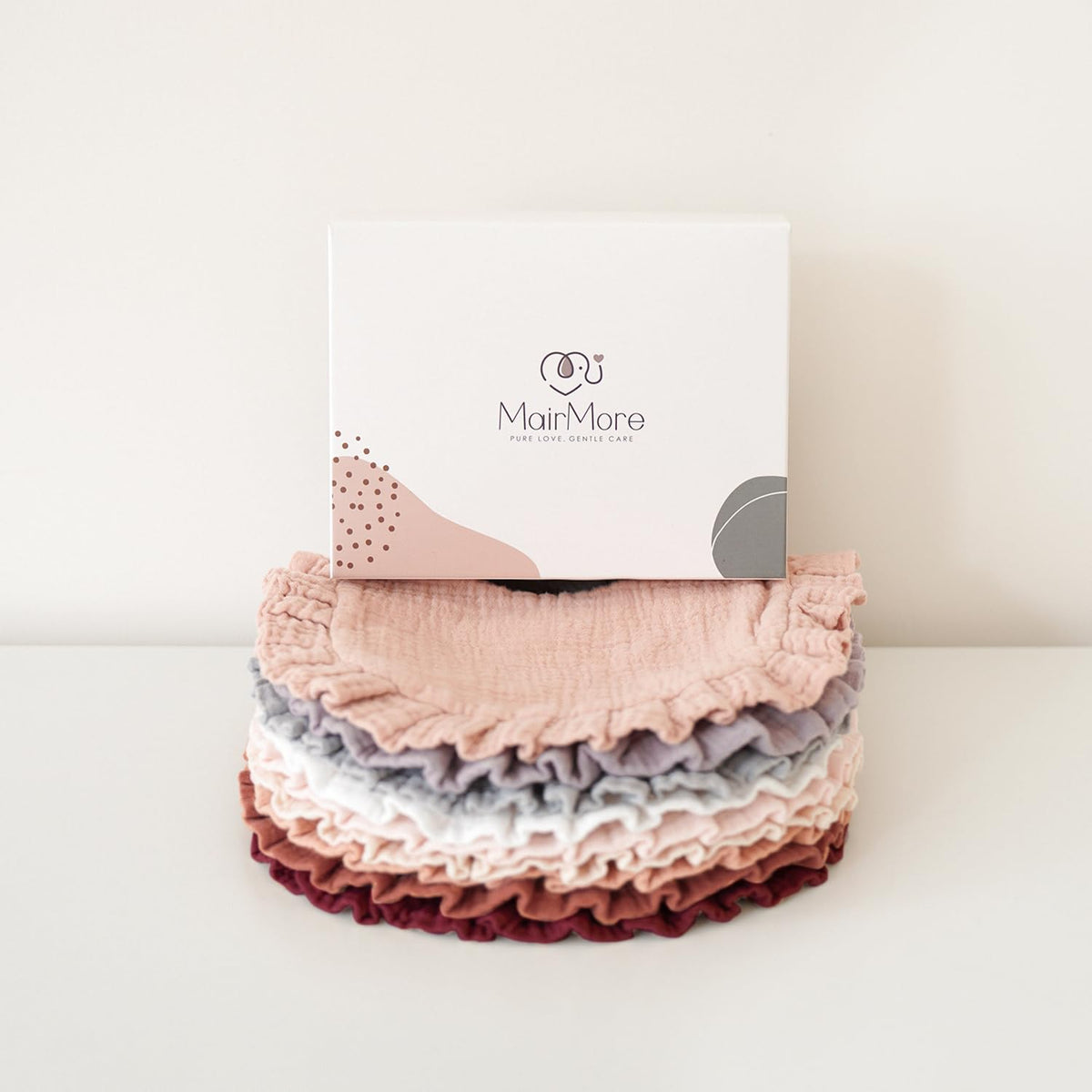 Stack of MairMore muslin bandana bibs in six colors -- blush pink, gray, lavender, white, dusty rose, and burgundy -- with textured ruffled edges, displayed in front of the branded white gift box.