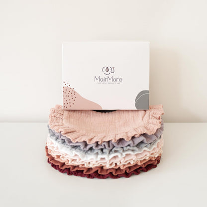 Stack of MairMore muslin bandana bibs in six colors -- blush pink, gray, lavender, white, dusty rose, and burgundy -- with textured ruffled edges, displayed in front of the branded white gift box.