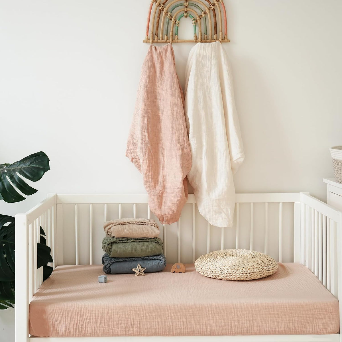 White wooden crib with a dusty pink muslin fitted sheet, topped with a stack of folded muslin blankets in blush, olive, and slate blue. A woven round cushion and small toys sit nearby. Two muslin swaddles hang above on a rainbow wall hook.