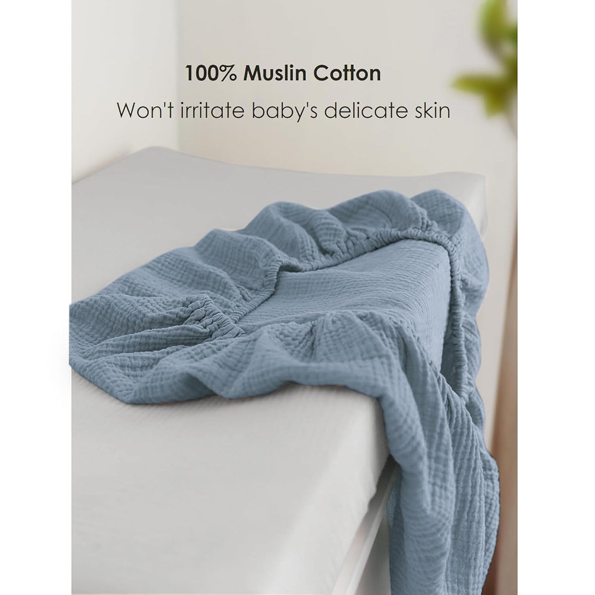 Dusty blue muslin cotton changing pad cover draped over a white changing pad, showing its textured, crinkled fabric and elasticized edges. Text reads '100% Muslin Cotton - Won't irritate baby's delicate skin.'