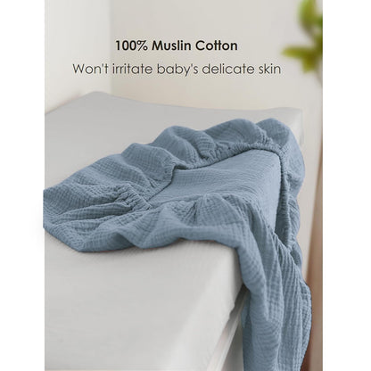 Dusty blue muslin cotton changing pad cover draped over a white changing pad, showing its textured, crinkled fabric and elasticized edges. Text reads '100% Muslin Cotton - Won't irritate baby's delicate skin.'