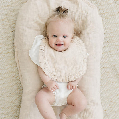 Smiling baby lying on a cream textured cushion wearing a light beige muslin bib with a ruffled edge trim over a white outfit. The bib has a soft, crinkled cotton texture and a wide coverage design.