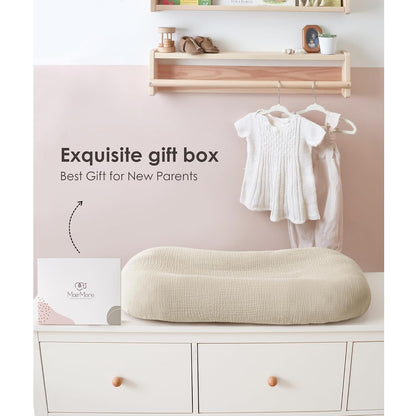 A cream-colored muslin baby lounger cover with a textured woven surface, displayed on a white dresser beside a branded gift box. Text reads 'Exquisite gift box - Best Gift for New Parents.' Nursery setting with shelf and hanging baby clothes.