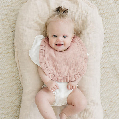Smiling baby lying on a cream muslin lounger wearing a dusty pink muslin bib with a ruffled edge trim. The bib has a soft, textured fabric appearance and a rounded shape covering the baby's chest.