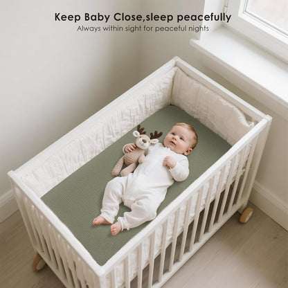 Baby lying on a sage green fitted sheet inside a white crib, wearing a white onesie and holding a knit reindeer toy. Text reads 'Keep Baby Close, sleep peacefully. Always within sight for peaceful nights.'