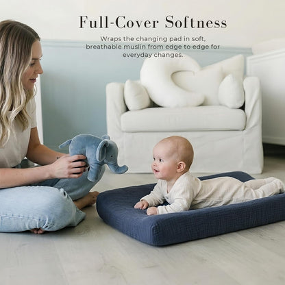 Baby lying on a navy blue muslin changing pad cover on a light wood floor, with a woman holding a blue elephant stuffed toy nearby. Text reads 'Full-Cover Softness' describing edge-to-edge breathable muslin coverage.