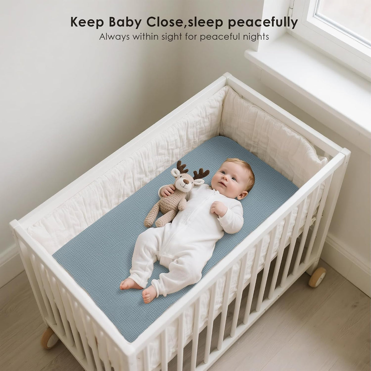 Baby lying on a blue fitted bassinet sheet inside a white wooden crib with bumper pads. The infant in a white onesie rests alongside a knit reindeer toy. Text reads 'Keep Baby Close, sleep peacefully.'