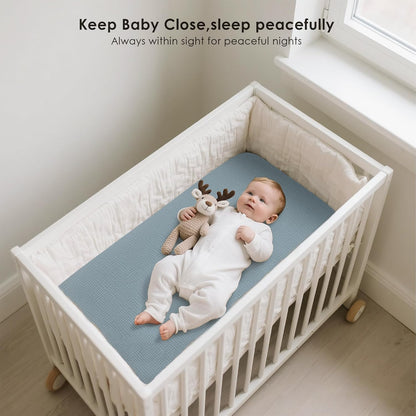 Baby lying on a blue fitted bassinet sheet inside a white wooden crib with bumper pads. The infant in a white onesie rests alongside a knit reindeer toy. Text reads 'Keep Baby Close, sleep peacefully.'