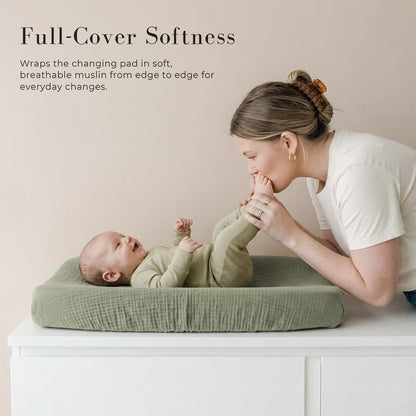 A baby lies on a sage green muslin changing pad cover atop a white dresser while a woman kisses the baby's foot. Text reads 'Full-Cover Softness' with a description of breathable muslin coverage edge to edge.