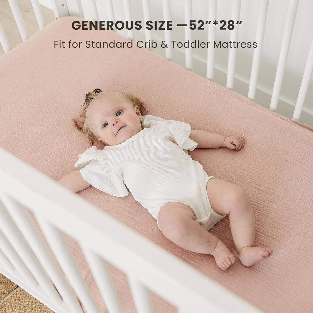 Baby lying on a dusty pink fitted crib sheet in a white crib. Text overlay reads 'Generous Size - 52"*28"' and 'Fit for Standard Crib & Toddler Mattress.'