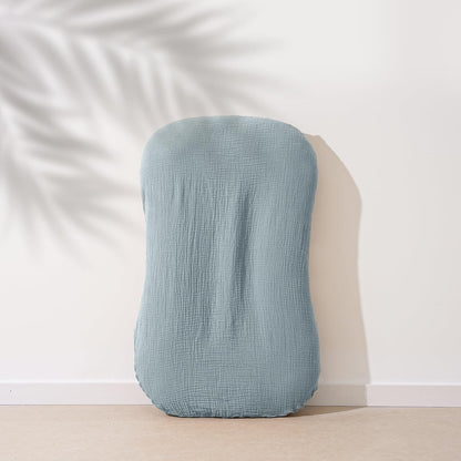 A dusty blue muslin baby lounger cover with a crinkled cotton texture, shaped to fit a curved infant lounger. The cover leans against a white wall with a soft palm leaf shadow in the background.