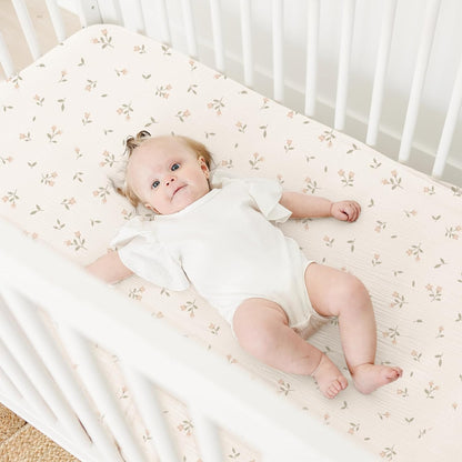 Baby lying on a cream fitted crib sheet with a small scattered floral pattern in soft pink and sage green. The sheet is shown on a standard white crib mattress, viewed from above at a slight angle.