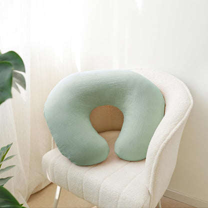 U-shaped nursing pillow cover in soft sage green muslin fabric, resting on a white textured armchair. The cover has a subtle woven texture and wraps a standard horseshoe-shaped nursing pillow.