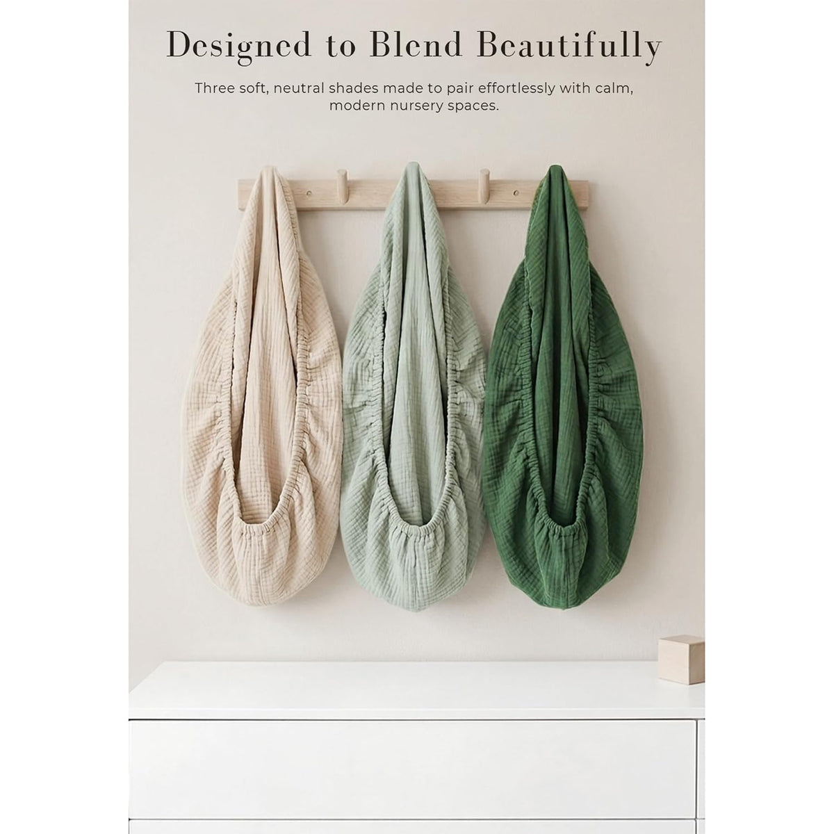 Three muslin cotton changing pad covers in beige, sage green, and forest green hang from a wooden wall hook rack. Each cover has a gathered, stretchy opening and textured woven fabric. Text reads 'Designed to Blend Beautifully.'