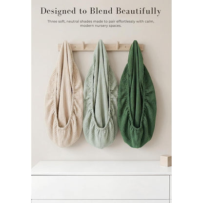 Three muslin cotton changing pad covers in beige, sage green, and forest green hang from a wooden wall hook rack. Each cover has a gathered, stretchy opening and textured woven fabric. Text reads 'Designed to Blend Beautifully.'