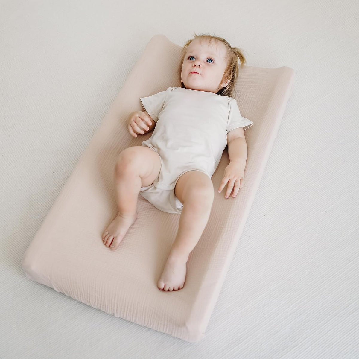 A baby lying on a changing pad covered with a soft clay-colored muslin cotton cover. The cover has a slightly textured surface and raised side edges, shown in a lifestyle context with the baby looking upward.