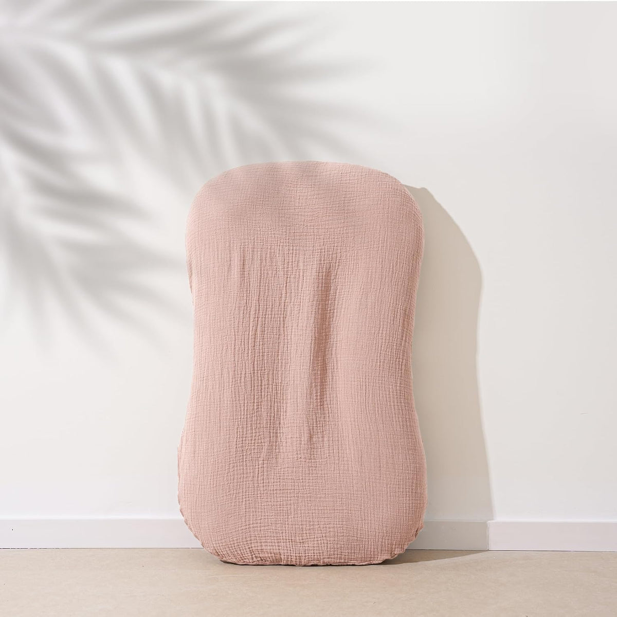 Dusty pink muslin cotton baby lounger cover with a crinkled texture, shaped to fit a curved infant lounger. Shown upright leaning against a white wall with a soft palm leaf shadow in the background.