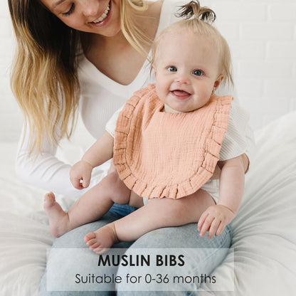 Smiling baby wearing a peach muslin bib with ruffled edges, seated on an adult's lap on a white surface. Text overlay reads 'MUSLIN BIBS - Suitable for 0-36 months.'