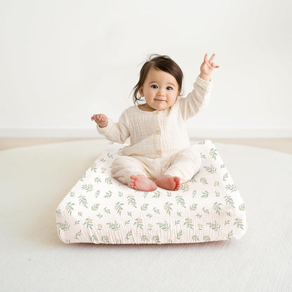 A smiling baby with one arm raised sits on a changing pad covered in a white muslin cover printed with small green leaf sprigs and tiny pink flowers. The textured fabric has a soft, gauzy appearance.