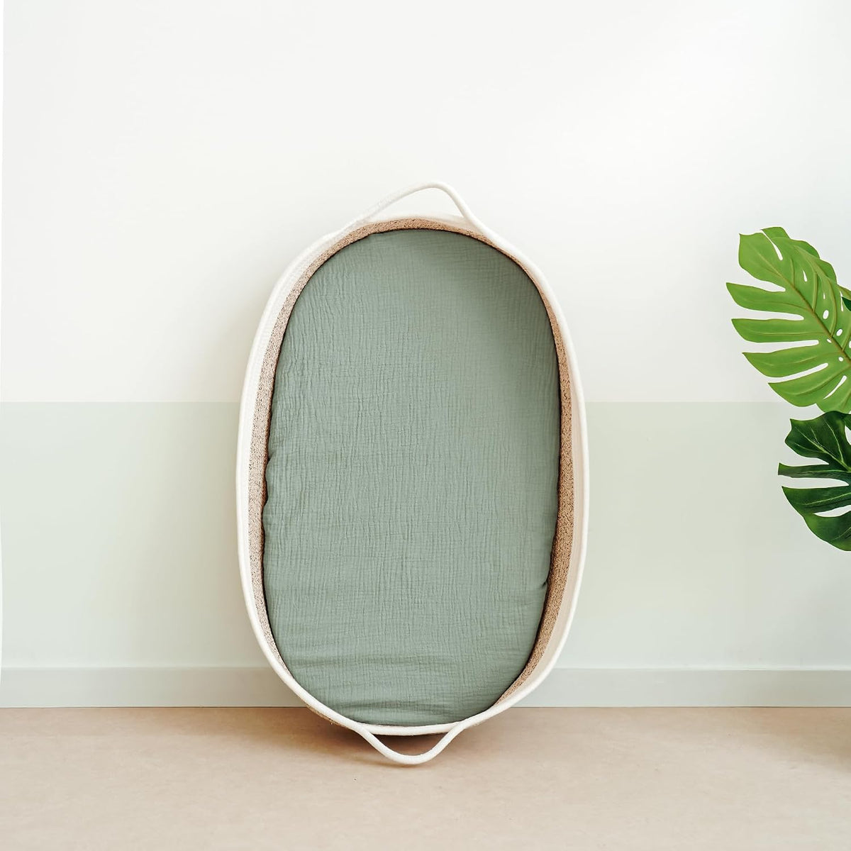 Oval bassinet with a sage green muslin fitted sheet, shown leaning upright against a two-tone white and pale green wall. The woven basket has cream-colored handles at each end. A monstera leaf is visible at right.