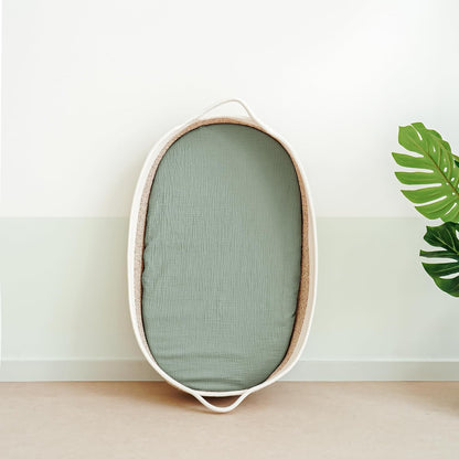 Oval bassinet with a sage green muslin fitted sheet, shown leaning upright against a two-tone white and pale green wall. The woven basket has cream-colored handles at each end. A monstera leaf is visible at right.