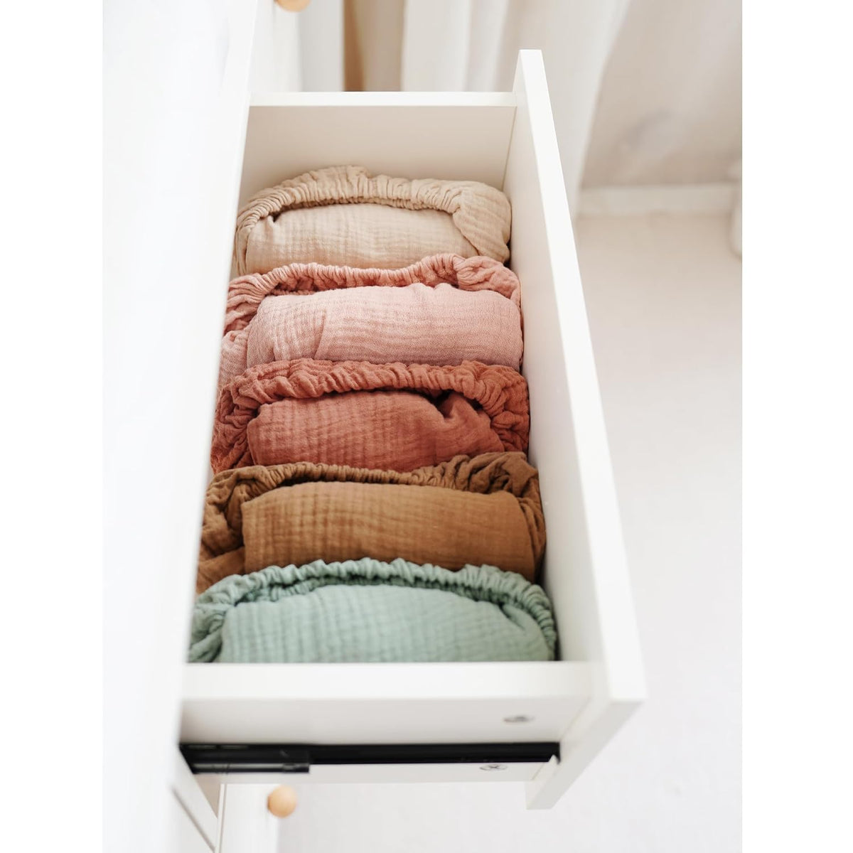 An open white drawer neatly organized with six rolled muslin changing pad covers in a gradient of colors: tan, dusty rose, pink, terracotta, golden brown, and sage green, showcasing the textured cotton fabric.