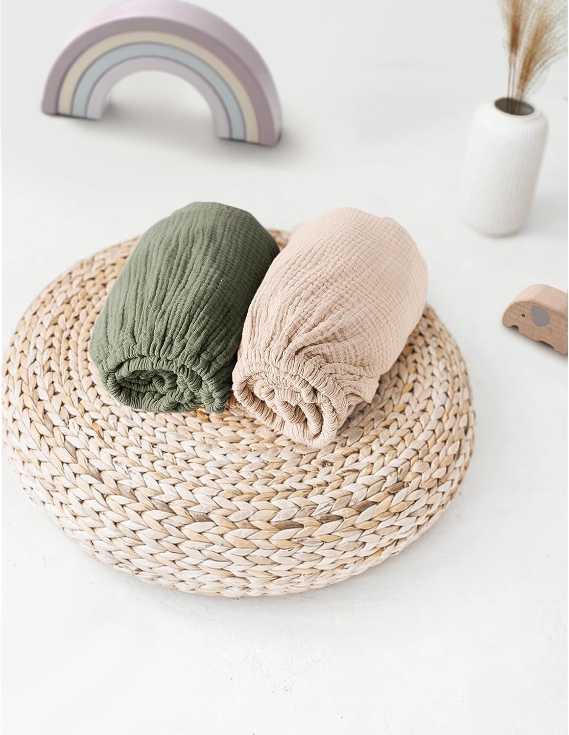 Two rolled muslin fitted sheets displayed on a round woven rattan trivet. One sheet is olive green and the other is light beige, both showing a crinkled cotton texture with elasticated edges.