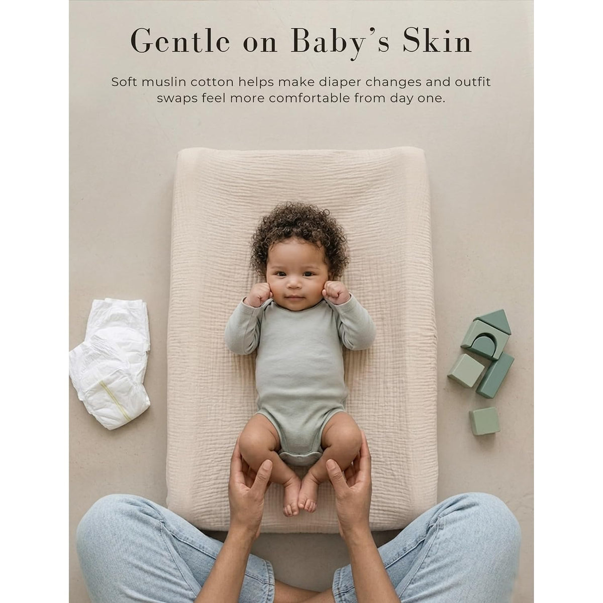 Promotional lifestyle image with text 'Gentle on Baby's Skin' showing a baby lying on a cream muslin changing pad cover. A caregiver's hands hold the baby's feet. A diaper and green toy blocks are visible nearby.