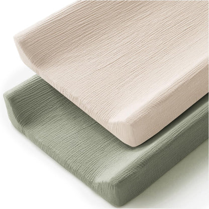 Two changing pad covers in a crinkled muslin fabric texture, one light blush pink and one sage green, stacked at an angle on a white background. Both covers wrap snugly around rectangular foam pads.