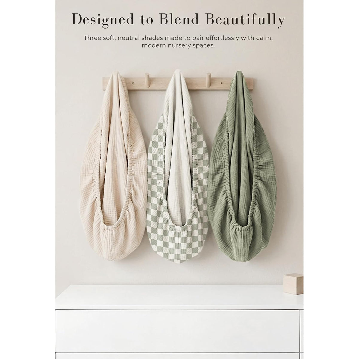 Three muslin changing pad covers hang on a wooden wall hook rack. Left is solid light beige, center has a sage green and white checkerboard pattern, right is solid sage green. All show gathered elastic edges and textured fabric.