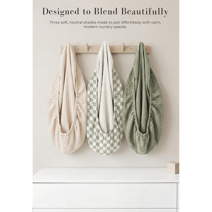 Three muslin changing pad covers hang on a wooden wall hook rack. Left is solid light beige, center has a sage green and white checkerboard pattern, right is solid sage green. All show gathered elastic edges and textured fabric.