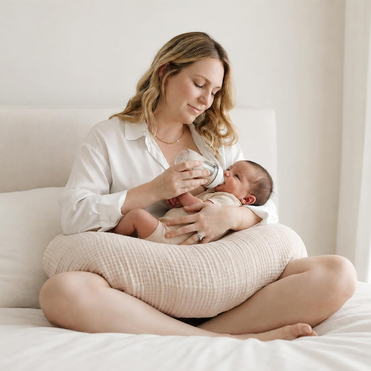 Woman sitting cross-legged on a white bed, bottle-feeding a newborn resting on a light beige textured nursing pillow wrap around her waist. The pillow has a soft, woven muslin-style cover in a warm neutral tone.