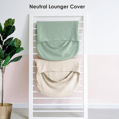 Two muslin lounger covers draped on a white slatted rack: one sage green and one ivory. Both show a textured cotton weave and curved pouch shape. Text above reads 'Neutral Lounger Cover'. A fiddle-leaf fig plant is visible to the left.