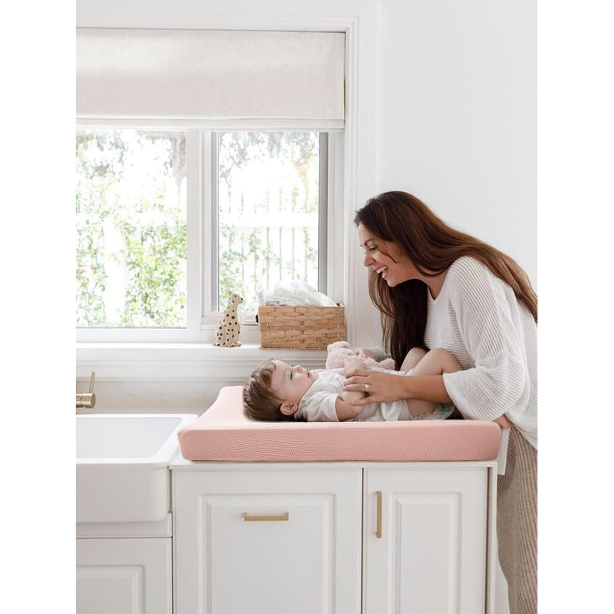 A smiling woman leans over a baby lying on a blush pink changing pad cover atop white nursery dresser. The textured cotton cover has a soft waffle or ribbed pattern in a warm pink tone.