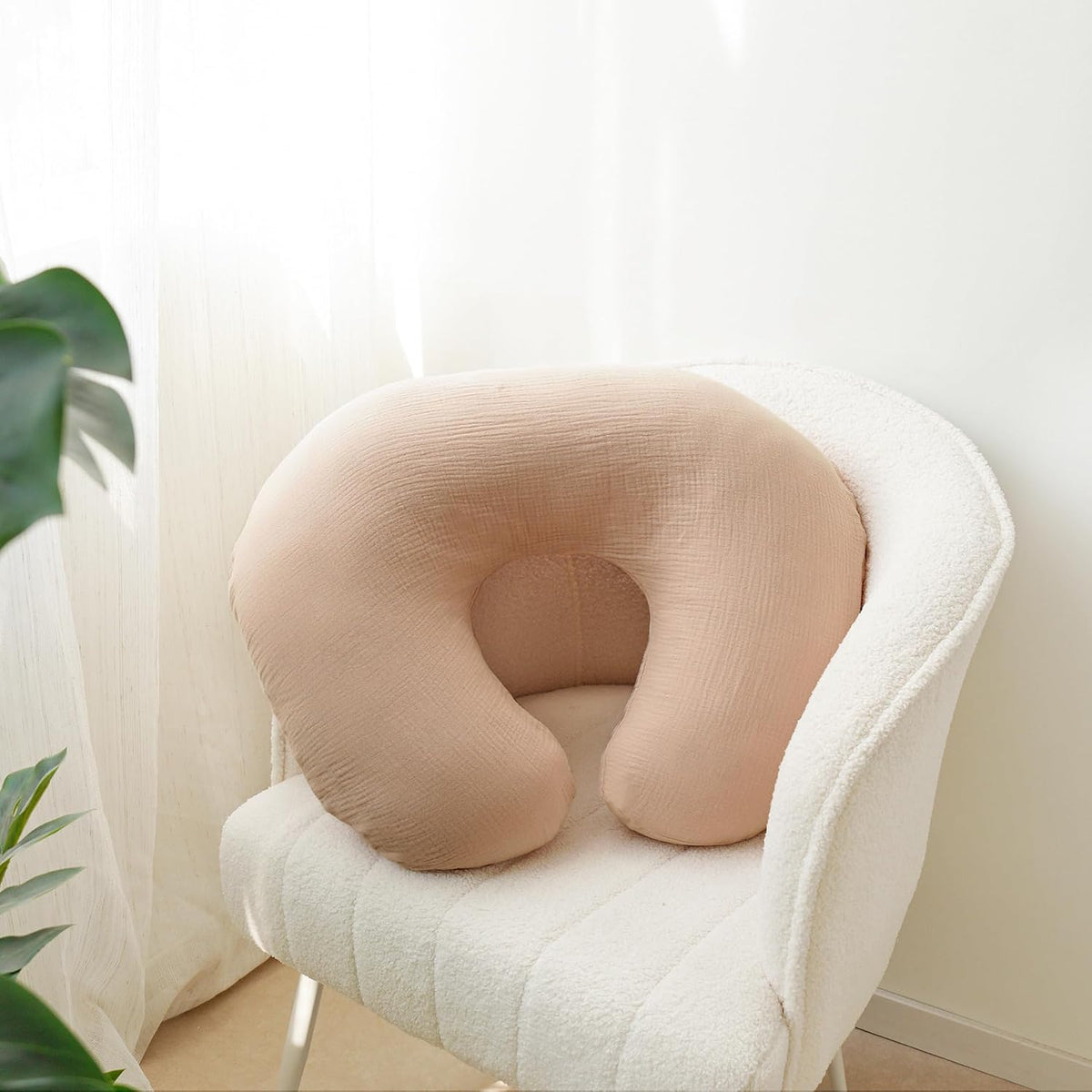 U-shaped nursing pillow in a soft beige muslin fabric cover, resting on a white textured armchair. The pillow has a curved horseshoe shape with a subtle woven texture and a small circular indent at the center opening.