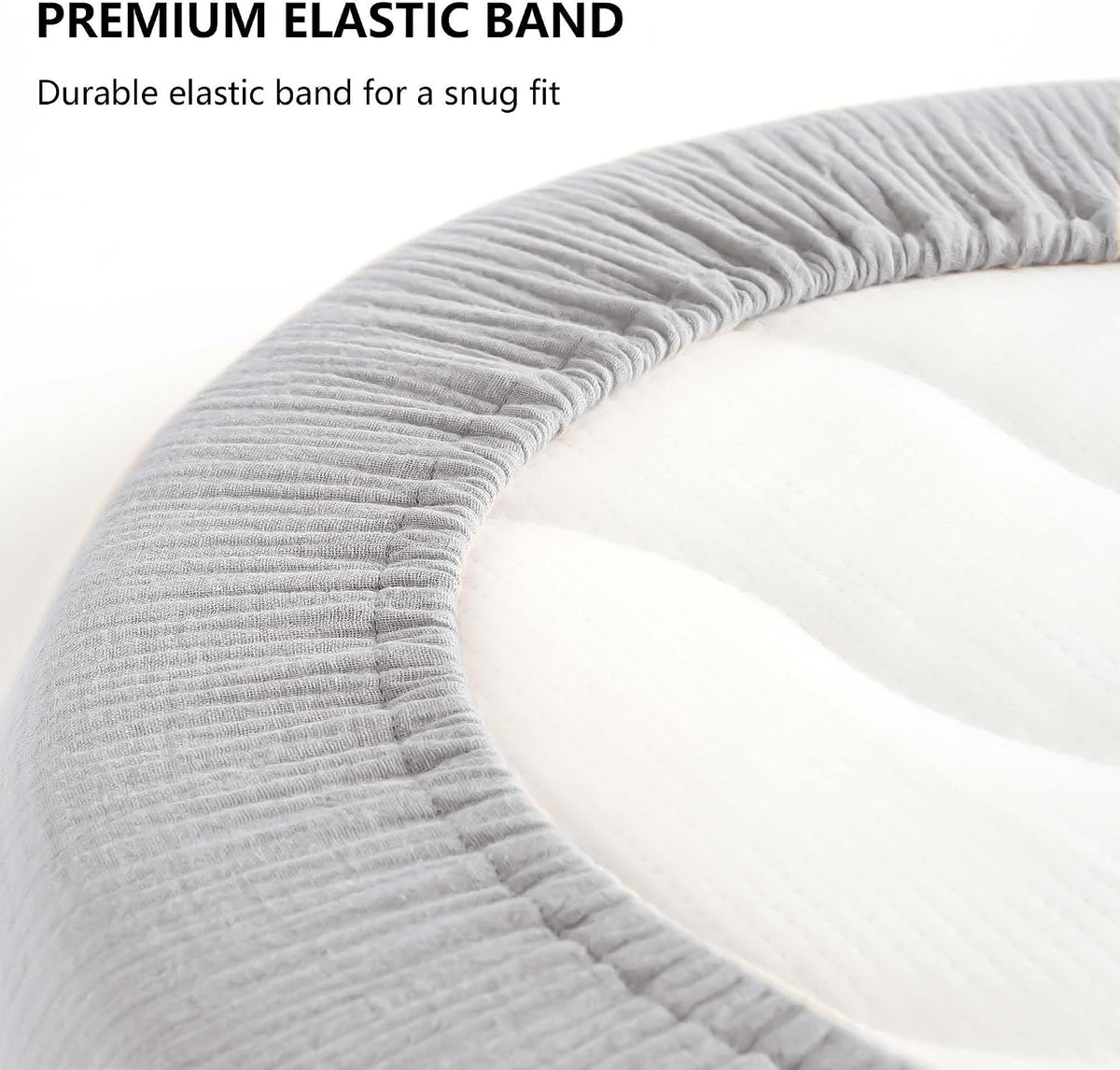 Close-up of a gray ribbed-texture fitted bassinet sheet stretched over a curved oval mattress edge, highlighting the elastic band. Text reads 'PREMIUM ELASTIC BAND - Durable elastic band for a snug fit.'