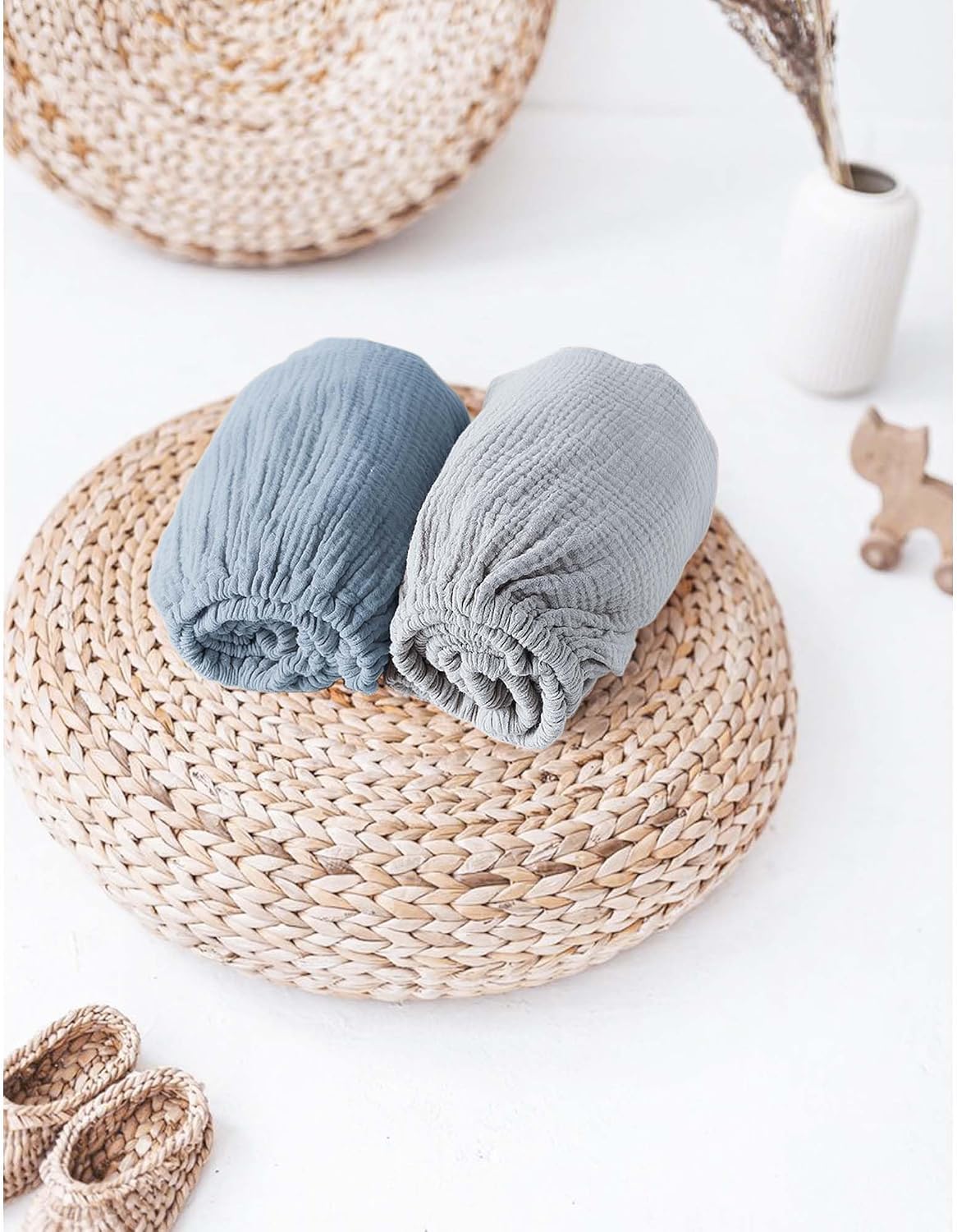 Two rolled muslin fitted sheets in dusty blue and light gray, placed side by side on a round woven rattan trivet. The crinkled cotton texture and elasticized edges are visible. Small knit booties and a wooden toy appear nearby.
