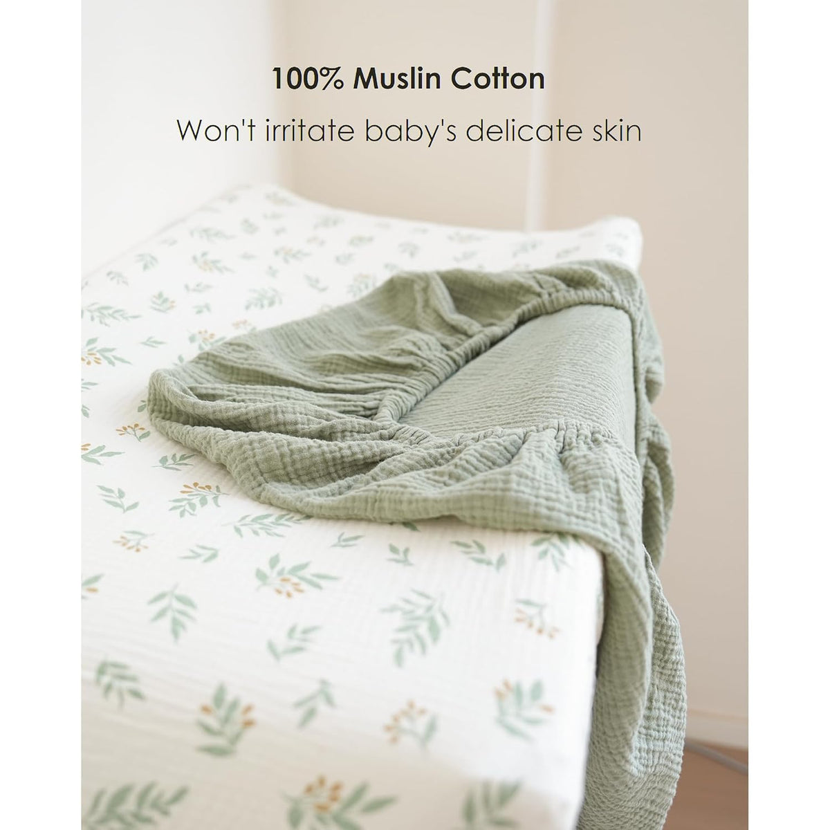Two muslin cotton changing pad covers on a changing table: one white with green leaf and orange berry print fitted on the pad, one solid sage green folded on top. Text reads '100% Muslin Cotton - Won't irritate baby's delicate skin.'