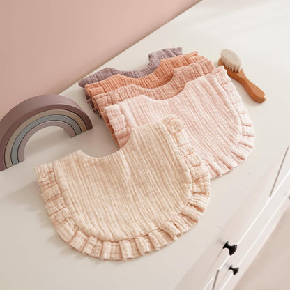 Four muslin baby bibs with ruffled edges fanned out on a white dresser, in graduating colors: cream, blush pink, peach, and dusty purple. A small rainbow toy and wooden brush are visible nearby.