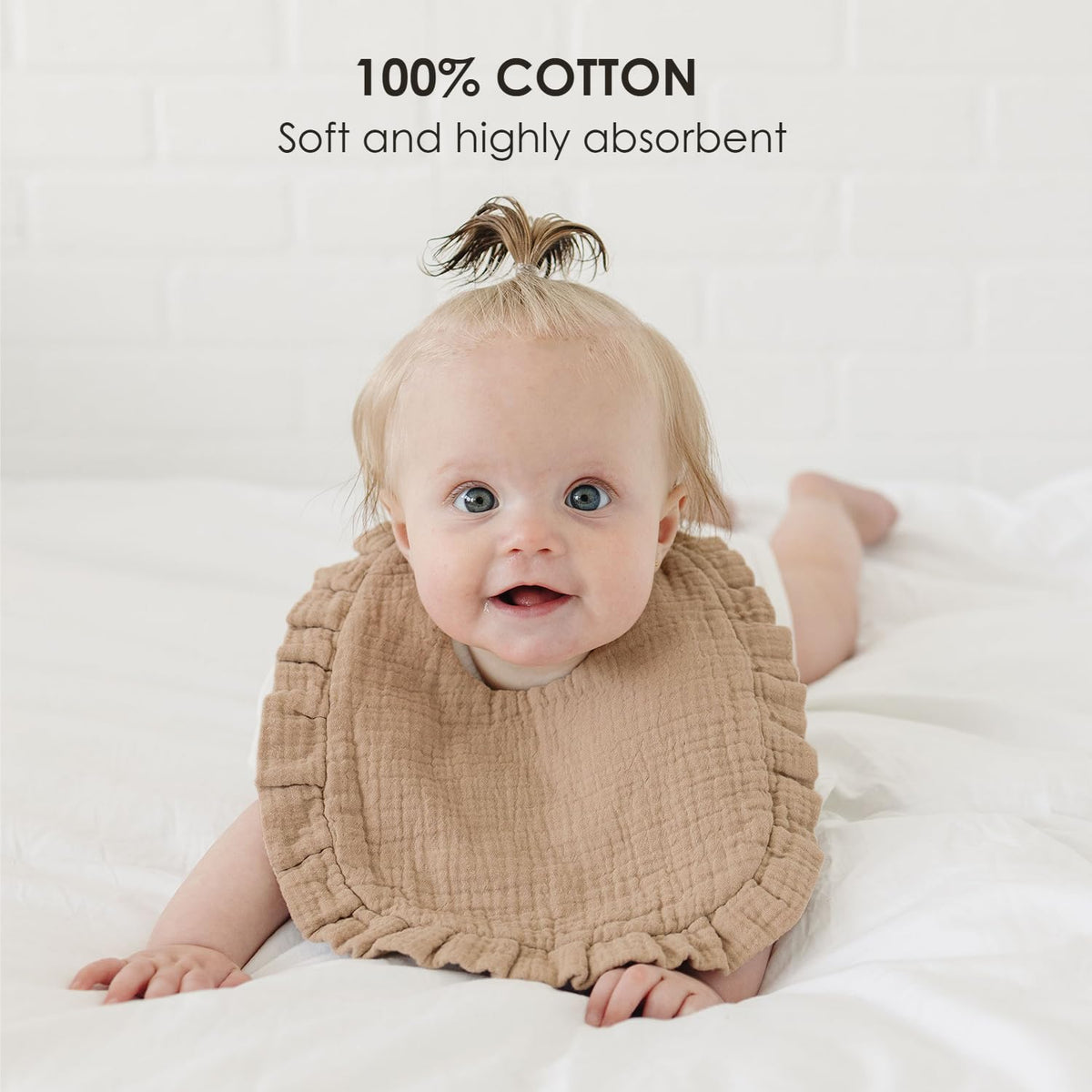 Smiling baby wearing a tan muslin cotton bib with ruffled edges, lying on a white surface. Text overlay reads '100% COTTON - Soft and highly absorbent.'