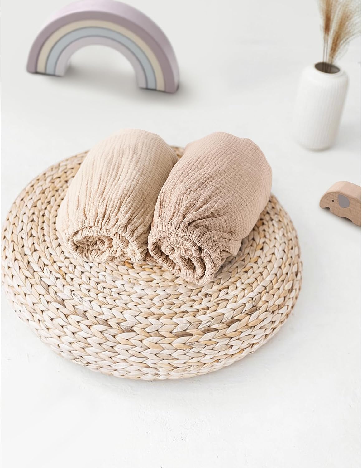 Two rolled muslin fitted bassinet sheets in light beige and slightly darker taupe tones, resting on a round woven rattan trivet. A pastel rainbow toy and small white vase with dried grass appear in the background.