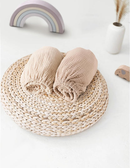 Two rolled muslin fitted bassinet sheets in light beige and slightly darker taupe tones, resting on a round woven rattan trivet. A pastel rainbow toy and small white vase with dried grass appear in the background.
