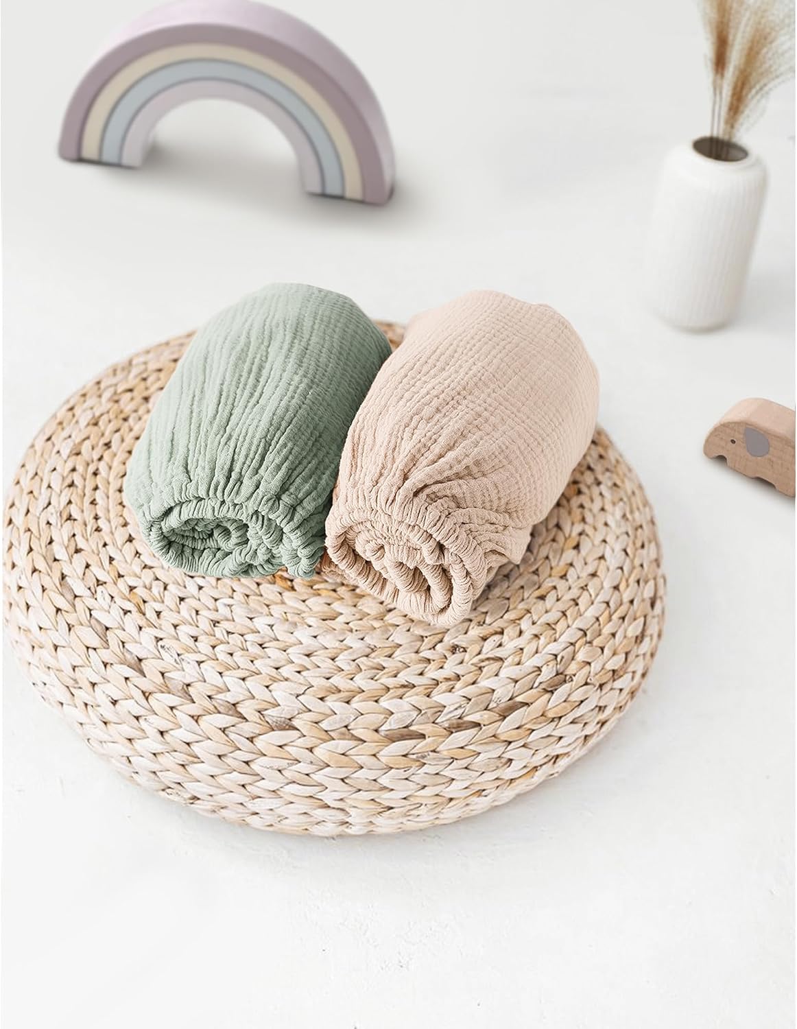 Two rolled muslin fitted bassinet sheets displayed on a round woven rattan trivet. One sheet is sage green and the other is light beige, both showing a crinkled cotton texture with elasticated edges.