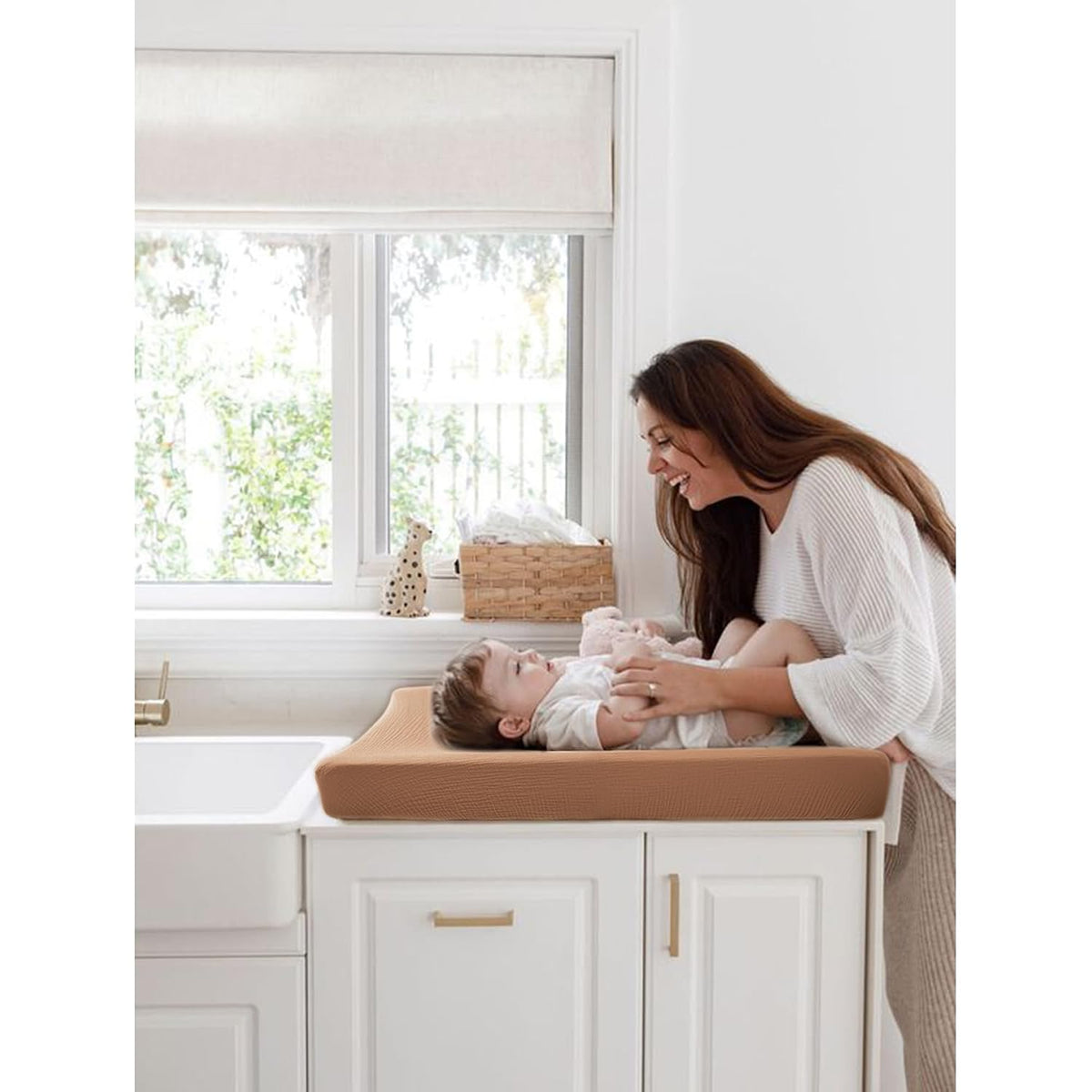 A smiling woman leans over a baby lying on a warm tan changing pad atop white cabinetry. The contoured changing pad has a textured, ribbed surface in a caramel-brown tone. A wicker basket and small decor sit on the windowsill behind.
