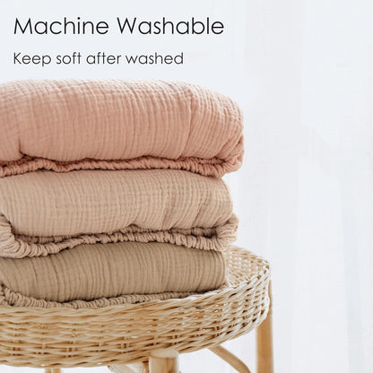Three folded muslin cotton blankets stacked on a wicker stool, in dusty rose, light tan, and khaki. Crinkled texture visible. Text reads 'Machine Washable - Keep soft after washed.'